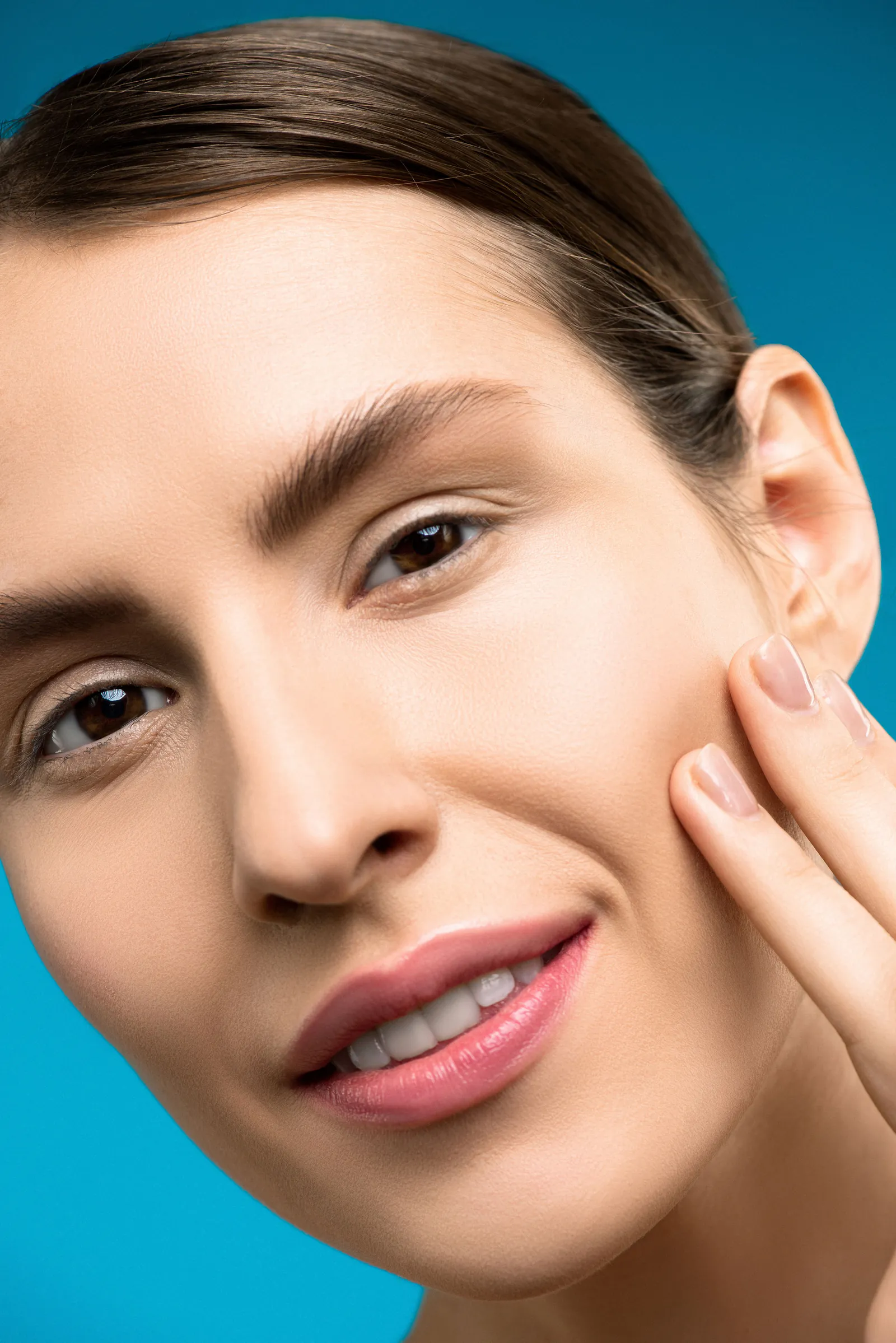 Close-up portrait of a smiling woman with smooth skin touching her cheek against a blue background.