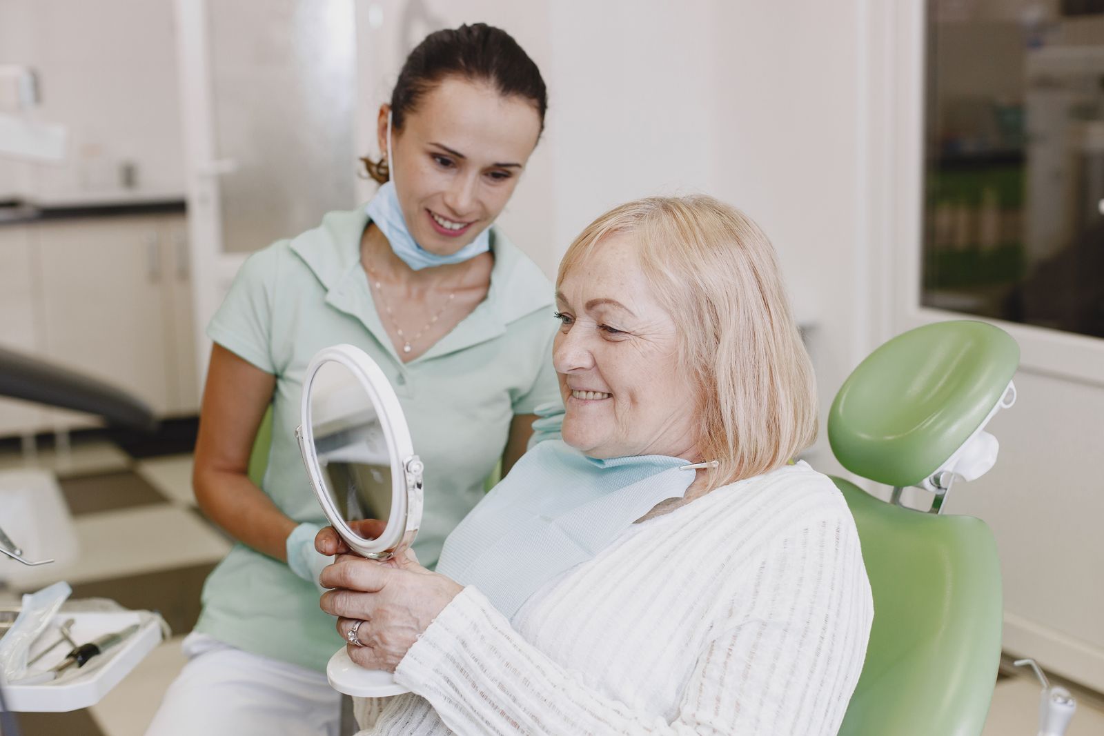 Smiling dental patient viewing her teeth in a mirror beside a clinician.